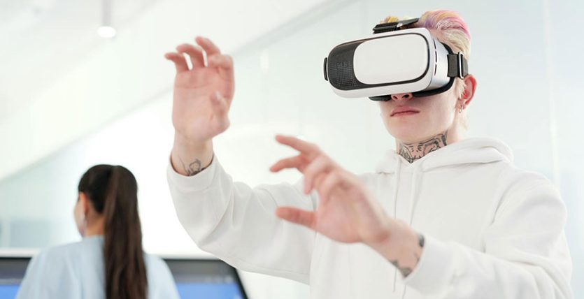 man wearing white hoodie using a VR goggles and a woman in front of a big screen in the background